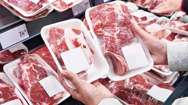 Shopper comparing packaged fresh beef cuts in a supermarket meat display, illustrating the crossfire of rising agricultural production costs, meat prices and growing food affordability pressures facing South African consumers in 2027.